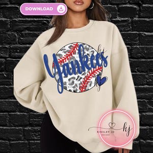 May include: Cream-colored sweatshirt featuring a baseball-themed design. The design includes the word "Yankees" in blue script, a baseball with red stitching, and a leopard print background. A blue heart is also included.