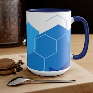 Pode incluir: Uma caneca de cerâmica branca com um padrão geométrico azul e branco. A caneca tem uma borda e uma alça azul marinho. A caneca está sobre uma superfície de madeira com grãos de café e um macaron de chocolate.