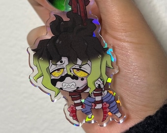 Puede incluir: Un llavero acrílico holográfico con un personaje de dibujos animados con piel verde, cabello negro y ojos amarillos. El llavero tiene un cierre en forma de corazón de oro rosa y un cierre de mosquetón. El personaje tiene un aspecto angustiado y viste un atuendo rojo y azul.
