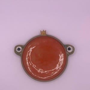Può includere: Un piatto in ceramica fatto a mano con un interno arancione vibrante e un bordo verde chiaro. Il piatto ha due piccole maniglie a forma di occhio e un accento a forma di corona dorata. Il design generale è circolare.