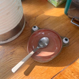 Può includere: Un poggiacucchiaio in ceramica fatto a mano a forma di rana, con un piccolo cucchiaio in acciaio inossidabile. Il poggiacucchiaio è marrone-rossastro con occhi verdi. Il cucchiaio ha un manico bianco.