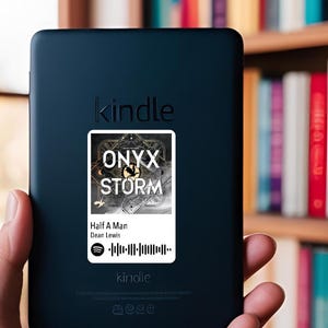 Puede incluir: Un lector de libros electrónicos Kindle negro con una pegatina que muestra "ONYX STORM" y "Half A Man" de Dean Lewis. El lector de libros electrónicos se sostiene en una mano, con una estantería de fondo.