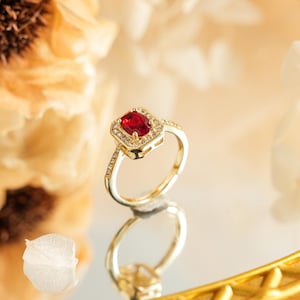 May include: A gold ring featuring a central oval-cut red gemstone surrounded by a halo of small clear stones. The band is thin and also accented with small clear stones along the sides, creating a bright and reflective appearance.