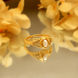 Op de afbeelding: Een gouden ring met een witte parel gezet in een decoratief ontwerp. De ring wordt weerspiegeld in een spiegelend oppervlak.