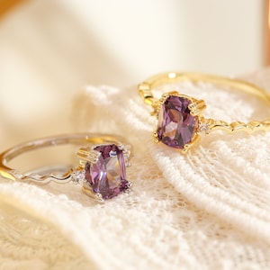 Peut inclure: Deux bagues avec des pierres précieuses violettes et de petites pierres claires sur un fond de dentelle blanche.