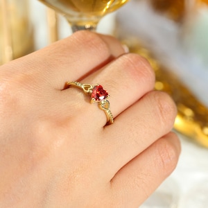 Könnte beinhalten: Ein goldener Ring mit einem roten herzförmigen Edelstein. Der Ring ist mit kleinen klaren Steinen auf der Band besetzt.