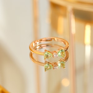 Può includere: Un anello d'oro con un design a forma di fiocco con due pietre preziose verdi e una pietra preziosa trasparente più piccola al centro.