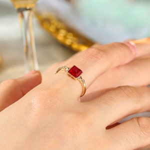 Può includere: Un anello d'oro con una pietra preziosa rossa quadrata e piccole pietre preziose chiare sulla fascia.