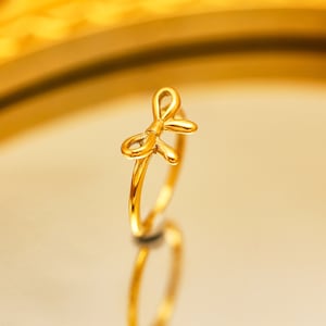 May include: A gold-colored ring with a bow design. The ring is on a gold-colored surface.