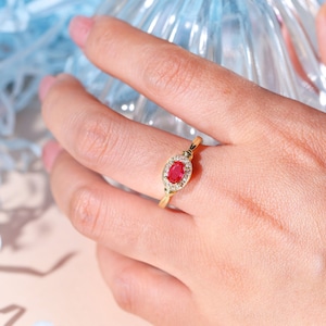 May include: A gold ring with an oval, bright red gemstone, encircled by smaller, clear stones. The ring is set on a slender gold band. The ring is shown on a hand, against a soft, neutral backdrop.