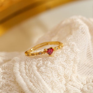 May include: A gold ring with a red heart-shaped stone and small clear stones around the band.