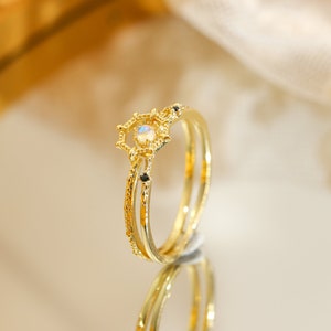 May include: A gold ring with a delicate, ornate design featuring a moonstone centre and black accents. The ring is layered with a second band, creating a unique and stylish look.