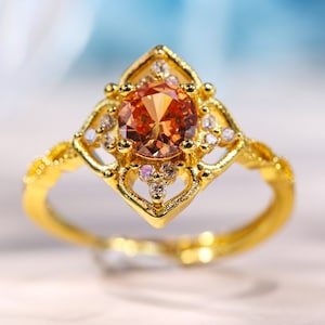 Könnte beinhalten: Ein goldener Ring mit einem großen, runden, orangefarbenen Edelstein, der von kleineren, klaren Edelsteinen umgeben ist. Der Ring hat ein filigranes, kompliziertes Design.
