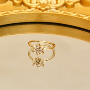 Può includere: Un anello d'oro con un designo a forma di fiore incastonato con pietre trasparenti. L'anello si riflette in uno specchio dorato.