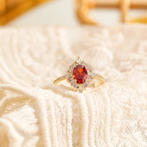 May include: A gold ring featuring a red oval gemstone surrounded by smaller clear gemstones. The ring is displayed on a white lace fabric.