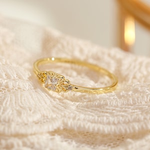 Könnte beinhalten: Ein zarter Goldring mit einem quadratischen, klaren Stein, der in einem floralen Design gefasst ist.