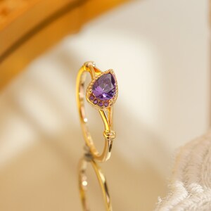 Op de afbeelding: Een gouden ring met een peervormige, dieppaarse edelsteen. De ring heeft een delicaat ontwerp met kleinere paarse stenen rond de hoofdsteen. De ring wordt gepresenteerd op een reflecterend oppervlak.