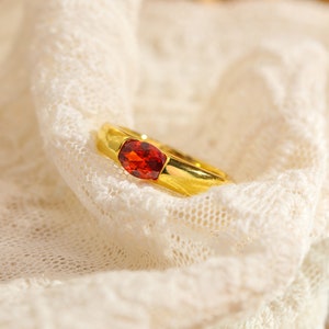 May include: A gold ring with a red gemstone set in the centre. The ring is on a white lace background.
