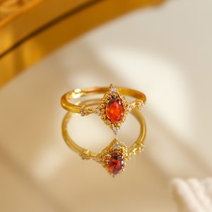 May include: A gold ring featuring a marquise-cut, red gemstone. The ring has an ornate design with small, clear accent stones. The ring is reflected on a shiny surface.