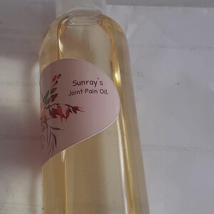 Puede incluir: Una botella de vidrio transparente con un líquido amarillo claro en su interior. La botella tiene una etiqueta blanca con el texto "Sunray's Joint Pain Oil" y un diseño floral.