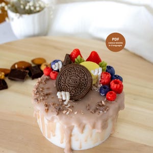 May include: A round candle shaped like a cake with light brown icing drips, topped with a dark brown Oreo cookie, red strawberries, blue blueberries, red raspberries, and a yellow macaron. A brown circular badge displays the text PDF CANDLE MAKING in 3 languages.