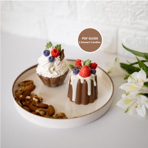 May include: Two dessert-shaped candles on a white plate with nuts. One candle is a cupcake with white frosting and berries. The other is a small cake with white icing and berries. A brown circle with the text "PDF GUIDE 2 Dessert Candles" is in the background.