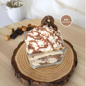 以下が含まれることがあります： クリーム、チョコレート、クッキーを重ねたデザートをテーマにしたキャンドル。正方形のガラス容器に入っています。キャンドルは木の輪切りに置かれ、円の中に「PDF CANDLE MAKING」という文字があります。
