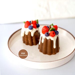 以下が含まれることがあります： チョコレート色のバントケーキ型のキャンドル2つ。白いフロスティングと赤いイチゴ、ブルーベリー、ラズベリーがトッピングされています。キャンドルは、まだらのオフホワイトのプレートに置かれています。画像には「PDF CANDLE MAKING」というテキストも含まれています。