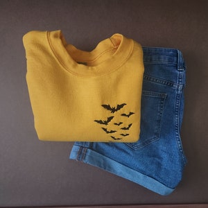 May include: A folded mustard yellow sweatshirt with a black bat graphic and blue denim shorts. The sweatshirt features a crew neck and a design of several bats in flight. The shorts are cuffed at the hem.