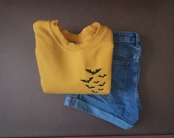 Peut inclure: Un sweat-shirt jaune moutarde plié avec un motif de chauve-souris noire et un short en jean bleu. Le sweat-shirt a un col rond et un motif de plusieurs chauves-souris en vol. Le short est retroussé.
