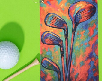 May include: A golf towel with a colorful abstract painting of golf clubs in shades of orange, blue, and purple. A white golf ball and a wooden tee are next to the towel on a green background.