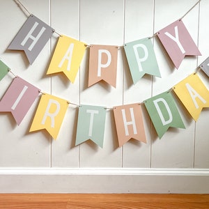 May include: A colorful "Happy Birthday" banner with pastel-colored pennants. Each pennant displays a letter in white, strung together with twine. The banner is hung against a white wall, creating a festive decoration.