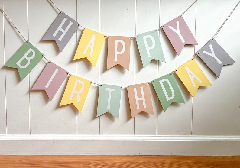 May include: A colorful "Happy Birthday" banner with pastel-colored pennants. Each pennant displays a letter in white, strung together with twine. The banner is hung against a white wall, creating a festive decoration.