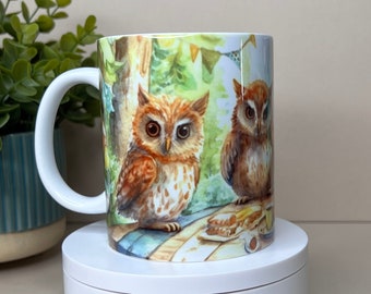 May include: A white ceramic mug with a colorful illustration of two owls sitting on a log in a forest setting. The owls are brown and white with large, round eyes. The mug has a handle and is decorated with green leaves and a blue sky.