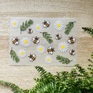 May include: A sheet of clear vinyl with white daisy flowers and brown bees with yellow stripes. The sheet is decorated with green leaves.