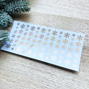 May include: A sheet of white sticker paper with a gradient of gold and white snowflake stickers. The stickers are arranged in rows and columns.