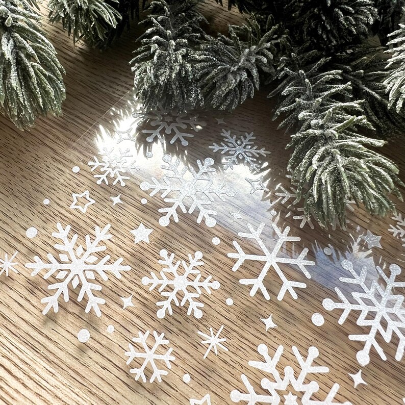 May include: A sheet of clear adhesive-backed vinyl with white glitter snowflake and star shapes. The sheet is on a wood surface.