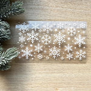 May include: A sheet of clear adhesive paper with white snowflake designs. The snowflakes are of various sizes and are scattered randomly across the sheet.