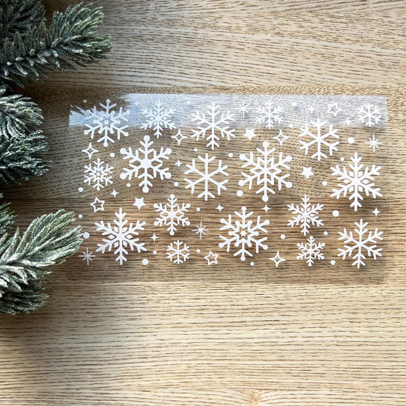 May include: A sheet of clear adhesive paper with white snowflake designs. The snowflakes are of various sizes and are scattered randomly across the sheet.