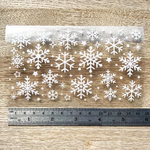 May include: A sheet of clear adhesive vinyl with white snowflake and star shapes. The sheet is approximately 6 inches wide and 10 inches long.