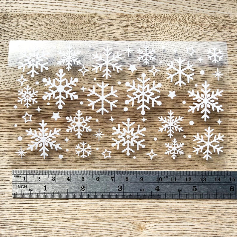 May include: A sheet of clear adhesive vinyl with white snowflake and star shapes. The sheet is approximately 6 inches wide and 10 inches long.