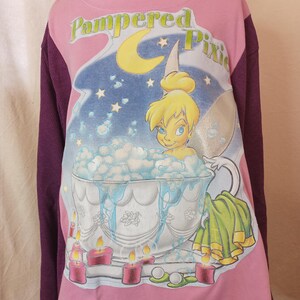 May include: Pink long-sleeved shirt with a purple sleeve, featuring a graphic of Tinkerbell in a bathtub with candles and the text "Pampered Pixie".