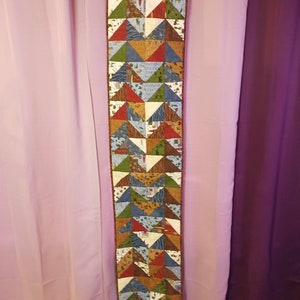 May include: A long, narrow table runner with a patchwork design of colorful triangles. The triangles are made of different fabrics with various patterns, including geometric shapes, floral designs, and woodland scenes. The runner has a brown border and is hanging on a wall.
