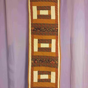 May include: A rectangular quilted table runner with a repeating pattern of orange, brown, and cream blocks. The design includes plaid and floral fabric sections. The runner is hanging against a purple curtain.
