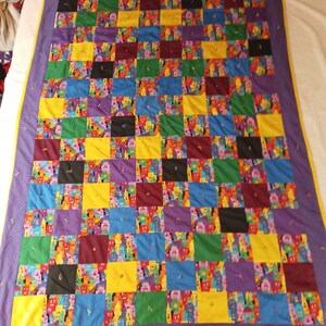 Puede incluir: Una colcha de retazos colorida con un patrón de cuadros. La colcha está hecha de varias telas en tonos de amarillo, morado, azul, verde, rojo y negro. La colcha está bordeada con un ribete amarillo.
