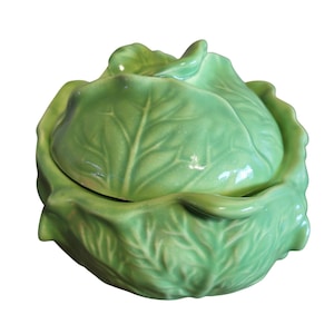 May include: A ceramic cabbage-shaped container with a lid. The container is a light green color and has detailed leaf textures. The lid fits snugly on top, creating a cohesive cabbage form. This decorative piece is likely for kitchen or dining use.