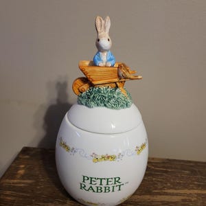 Pode incluir: Um pote de cerâmica branca Peter Rabbit com tampa. A tampa apresenta uma figura de Peter Rabbit em um carrinho de mão, com um pássaro pequeno. O pote tem um design floral e as palavras "PETER RABBIT" em verde.