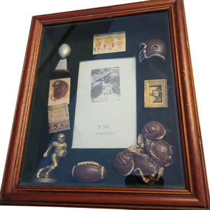 May include: A framed shadow box with football memorabilia. The frame is a dark wood color. Inside are miniature football items, including a helmet, football, and a small statue of a football player. A 4"x6" photo frame is in the center.