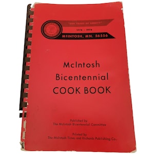 Può includere: Un libro di cucina rosso intitolato "McIntosh Bicentennial COOK BOOK". La copertina presenta testo e un emblema circolare con la scritta "200 YEARS OF LIBERTY" e le date 1776-1976. Il libro è rilegato a spirale.