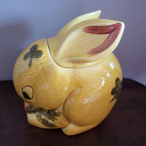 Może przedstawiać: Ceramiczny słoik na ciasteczka w kształcie królika w jasnożółtym kolorze. Uszy mają czerwonobrązowe wnętrze, a słoik jest ozdobiony zielonymi wzorami koniczyny. Królik jest w pozycji siedzącej, z głową obróconą na bok.